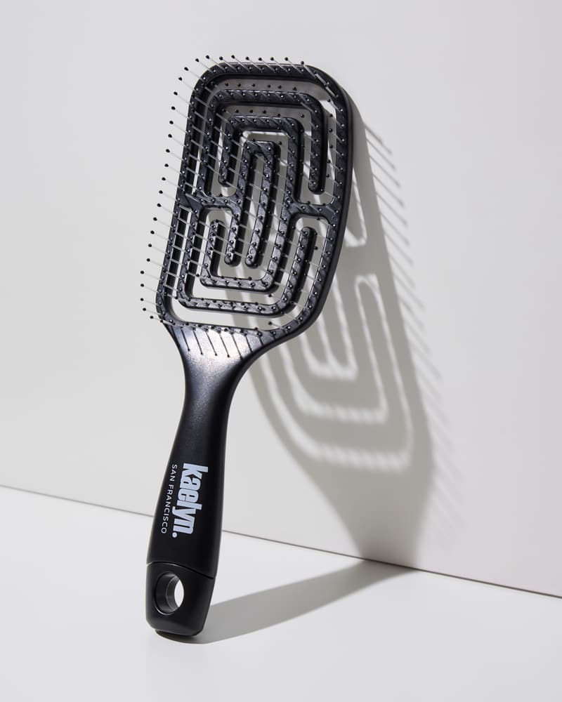 Black detangling brush with a maze-pattern vented head and a kaelyn. San Francisco logo on the handle, leaning against a light gray wall.