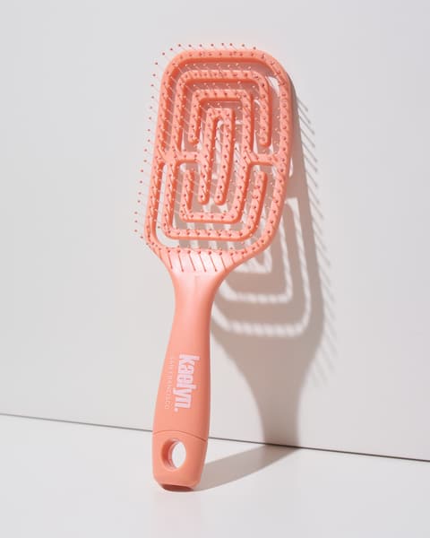 Coral detangling brush with a maze-pattern vented head and a hanging loop at the end of the handle, leaning against a light gray wall.