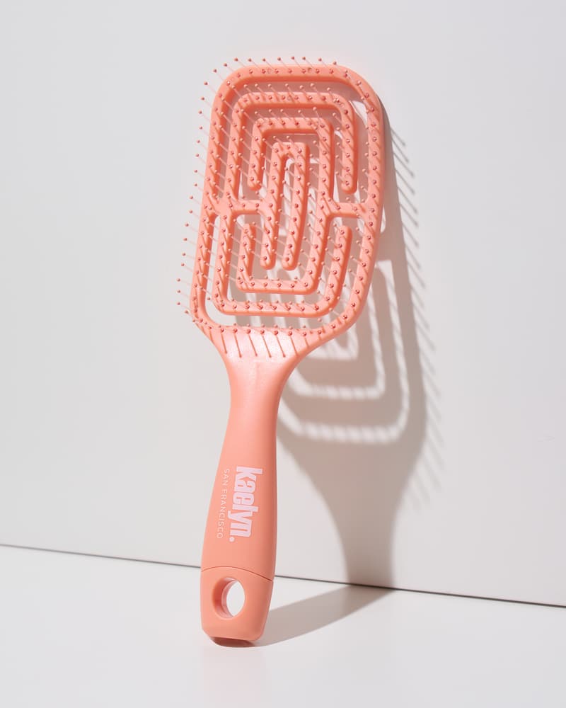 Coral detangling brush with a maze-pattern vented head and a hanging loop at the end of the handle, leaning against a light gray wall.