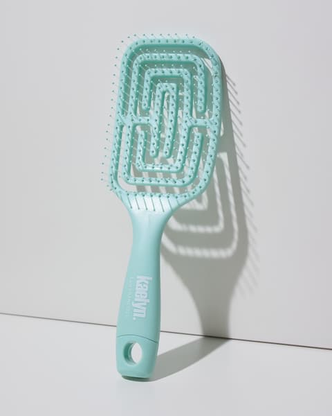 Mint detangling brush with a maze-pattern vented head and a hanging loop at the end of the handle, leaning against a light gray wall.