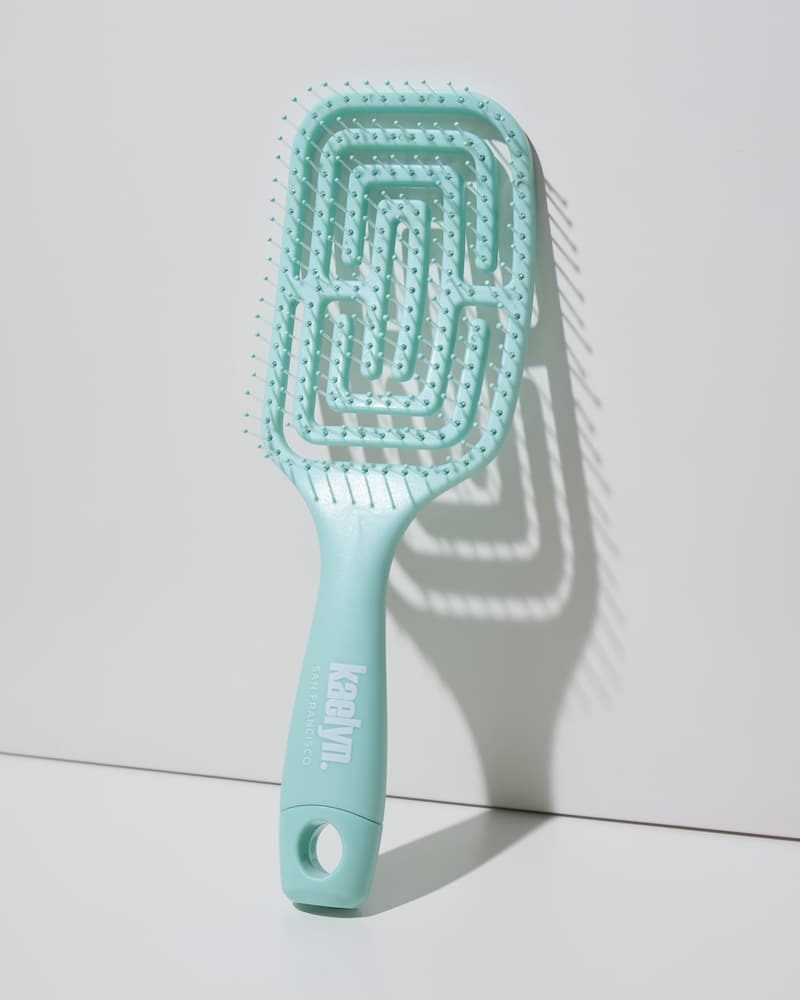 Mint detangling brush with a maze-pattern vented head and a hanging loop at the end of the handle, leaning against a light gray wall.