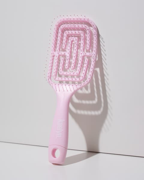 Light pink detangling brush with a maze-pattern vented head and a hanging loop at the end of the handle, leaning against a light gray wall.