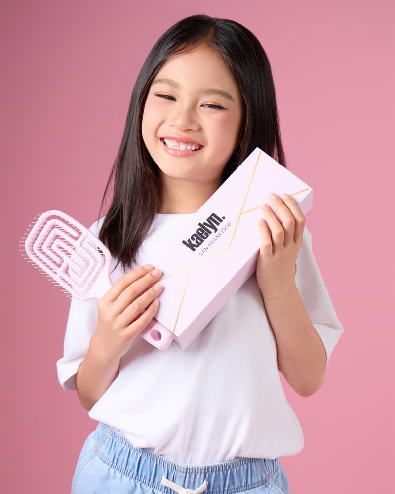 Smiling girl in a white kaelyn. t-shirt holding a pink hairbrush and a light pink box against a pink backdrop, cropped from head to waist.