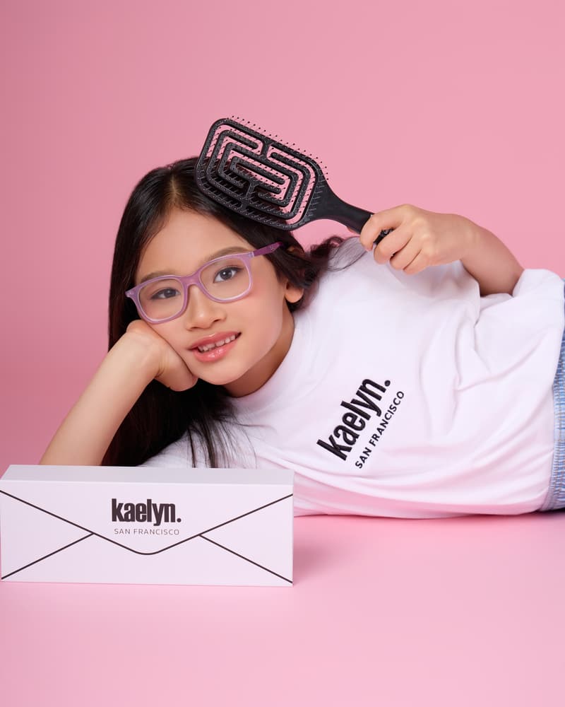 Girl in glasses lying on a pink floor with a black hairbrush in her hand and a white kaelyn. gift box beside her.
