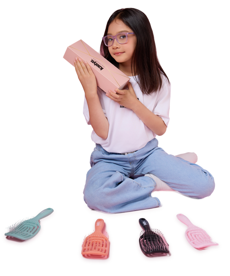 Girl in glasses sitting on a pink floor hugging a pink box, with four hairbrushes in mint, coral, black and pink on the floor in front of her.