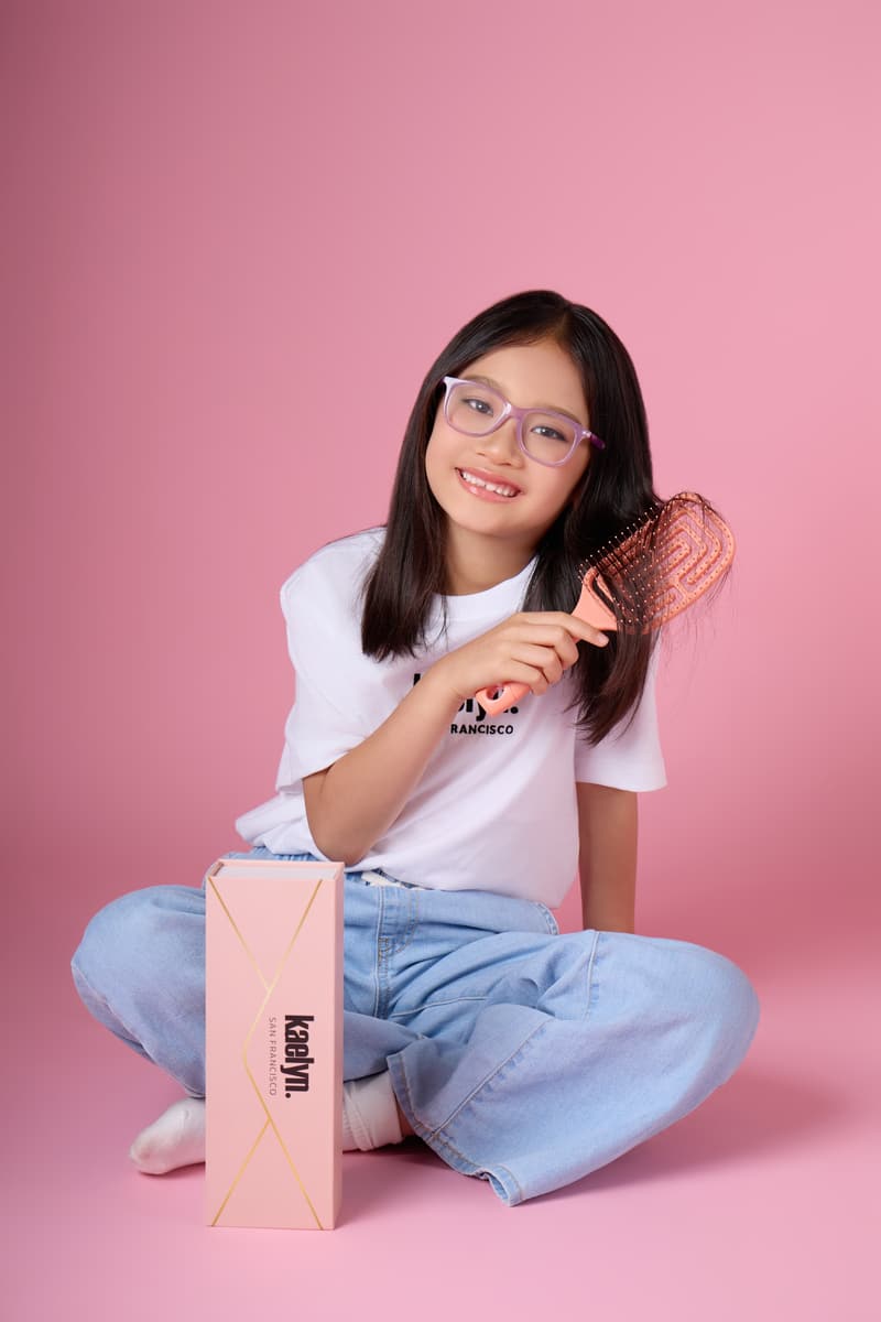 Girl in glasses sitting cross-legged beside a coral gift box, brushing her hair with a coral brush.