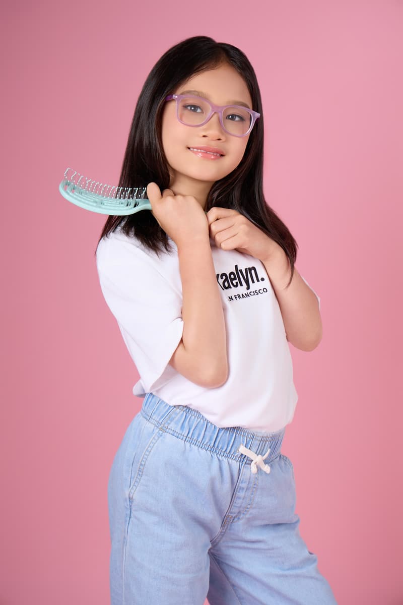 Girl in glasses holding a mint hairbrush up to her chin, wearing a white kaelyn. t-shirt and light jeans.