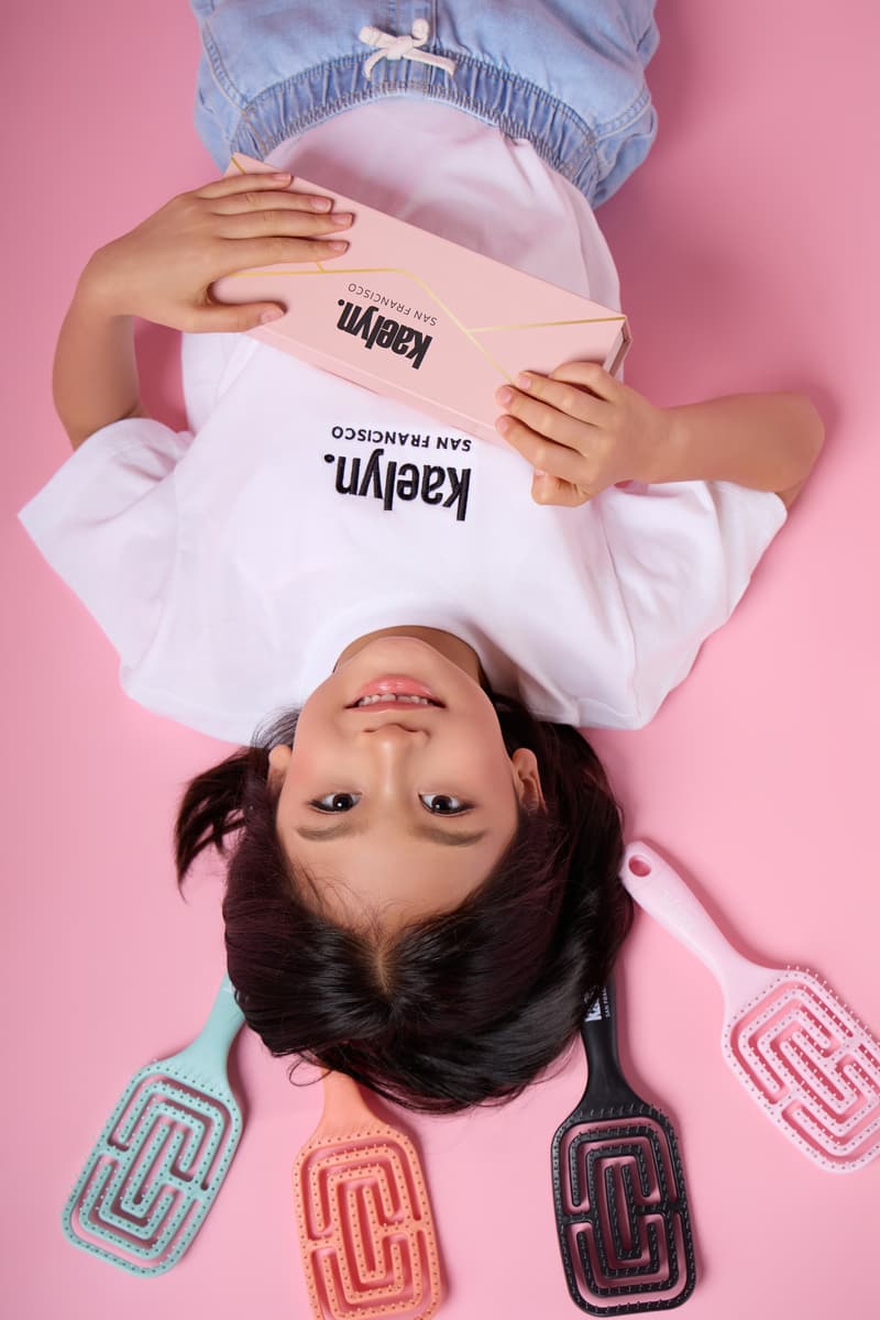 Top-down view of a girl lying on a pink floor, upside down, holding a light pink box, with four hairbrushes fanned out beside her head.