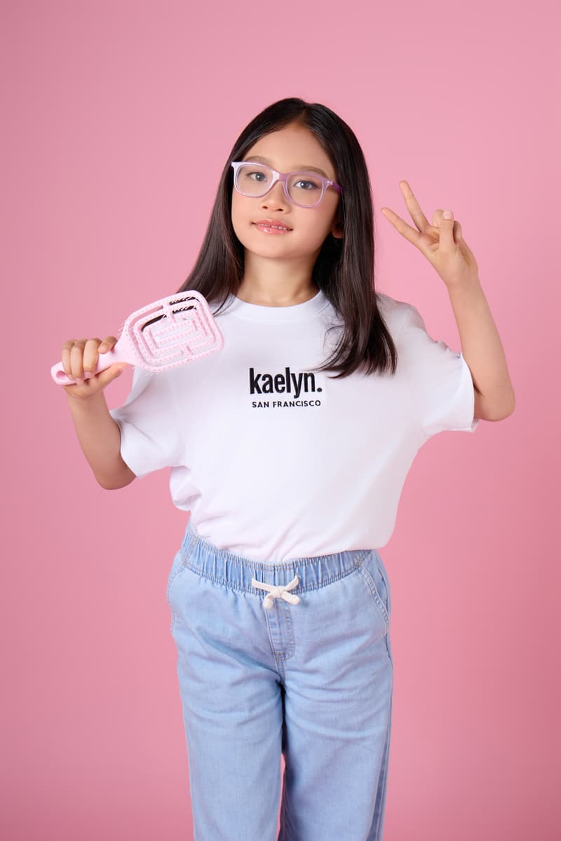 Girl in glasses holding a pink hairbrush in one hand and making a peace sign with the other, in a white kaelyn. t-shirt.