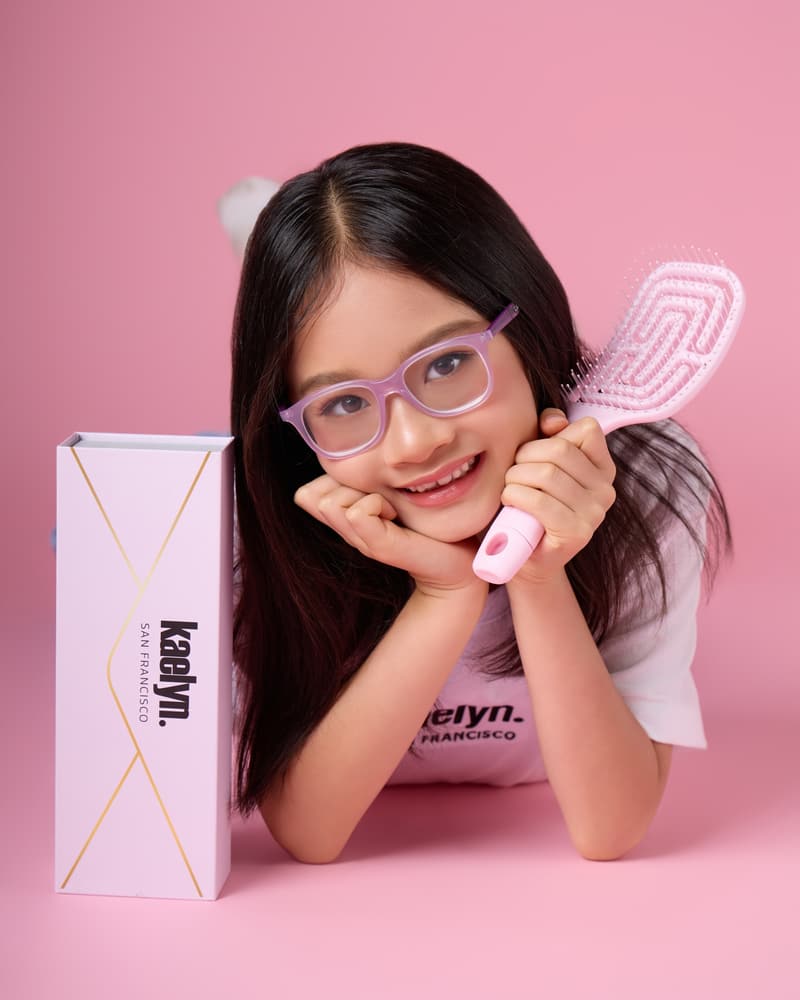 Girl in glasses lying on her front with her head on her hands, holding a pink hairbrush next to a pink kaelyn. box.
