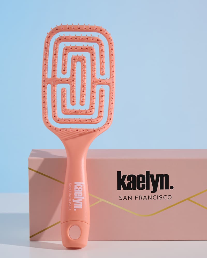 Coral brush standing in front of a peach kaelyn. San Francisco gift box with a thin gold line, on a light blue background.